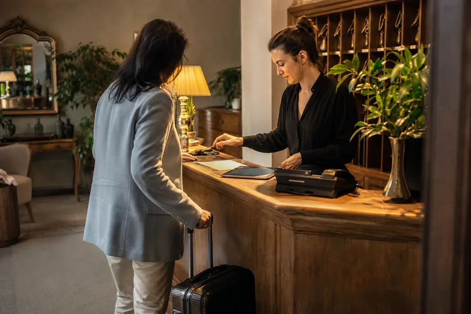 Recepcjonistka w małym hotelu obsługuje gościa z walizką, na ladzie tablet i telefon